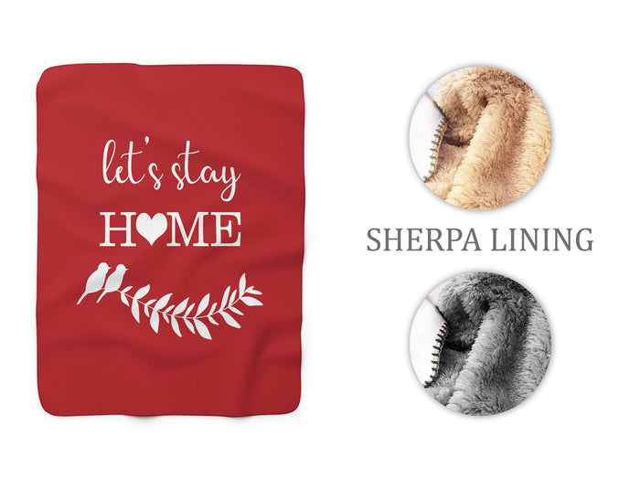 A red sherpa fleece throw blanket with the text 'Let's Stay Home' with a heart in place of the "o" and a white silhouette pair of love birds nestled among a branch with leaves on the front. A close-up of the fleece side shows a both brown and gray sherpa fleece textures.